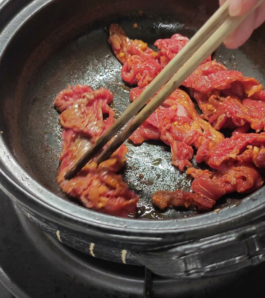 Geschnittenes Rindfleisch wird in einem dunklen Topf mit hölzernen Essstäbchen angebraten.