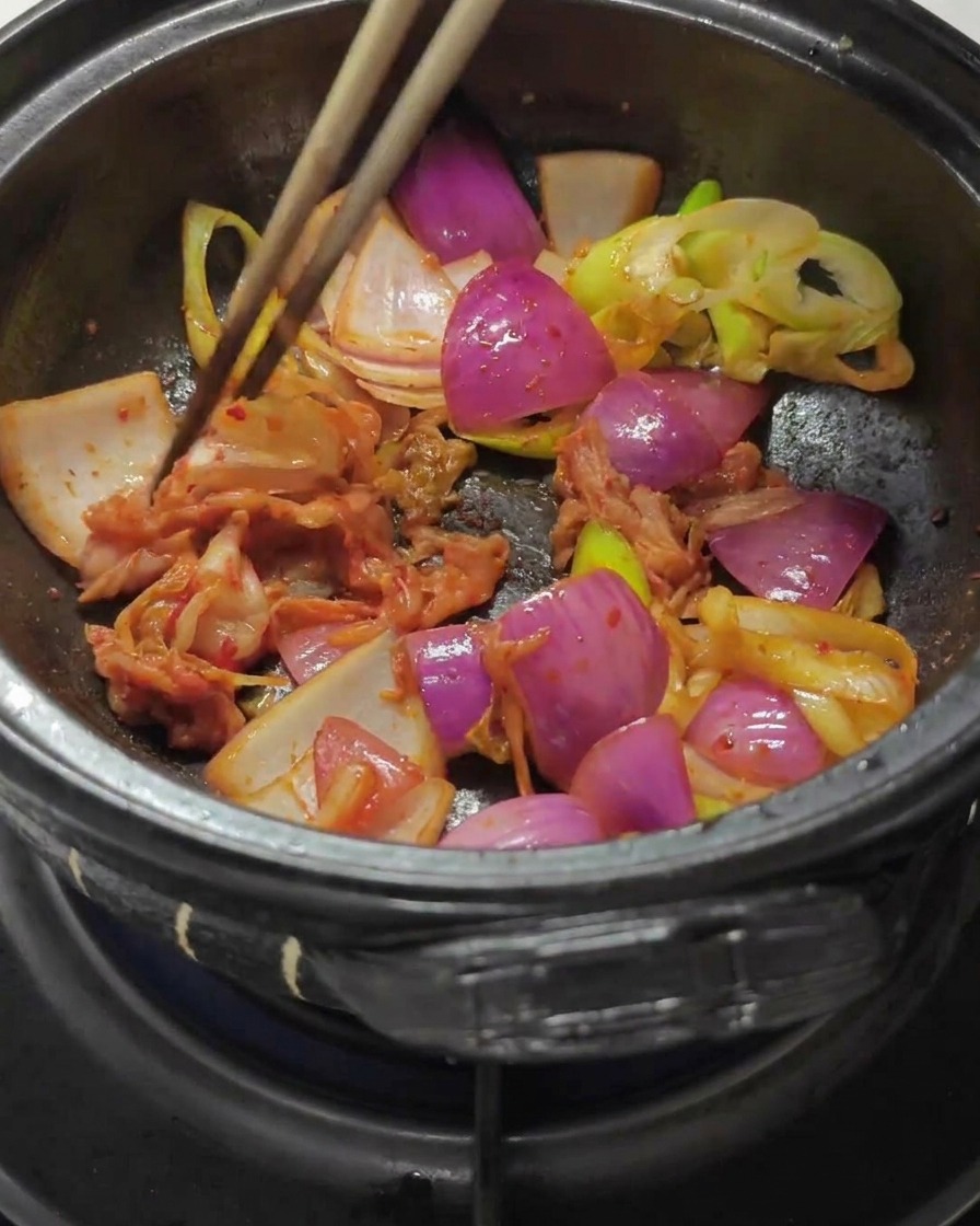 Essstäbchen mischen gehacktes Kohl-Kimchi und rote Chilipaste unter die angedünsteten Zwiebeln in einem Steintopf.