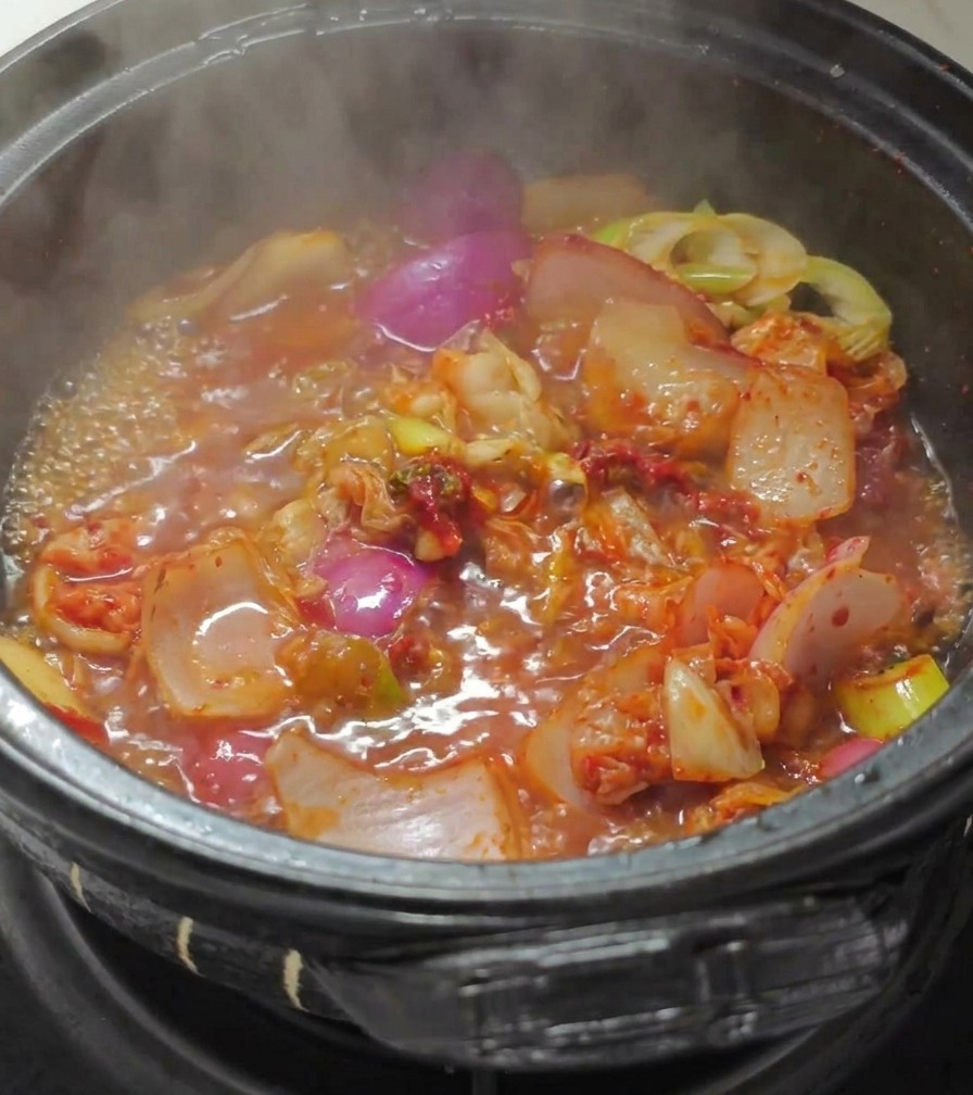 Kochendes Wasser wird in einen schwarzen Steintopf gegossen, der angebratenes Kimchi, rote Zwiebeln und Frühlingszwiebeln in einer kräftig roten Brühe enthält.