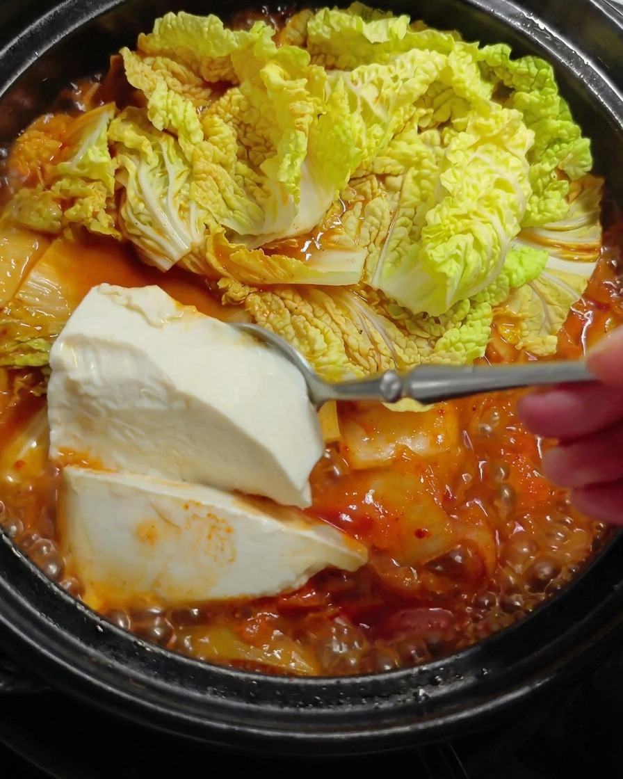 Ein großer Löffel mit weichem, weißem Tofu wird vorsichtig in einen heißen, brodelnden roten Eintopf mit Kohlblättern getaucht.