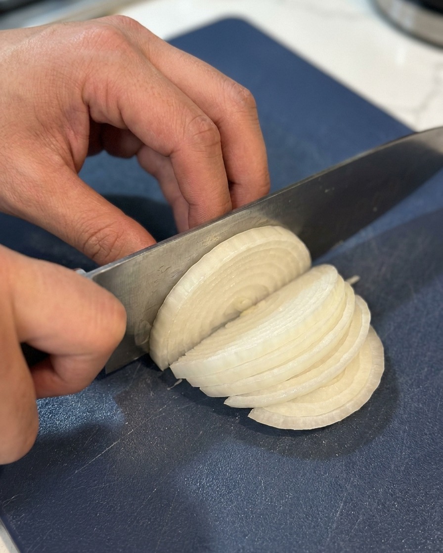 Hände schneiden eine halbierte weiße Zwiebel mit einem Kochmesser in feine Streifen auf einem blauen Schneidebrett.