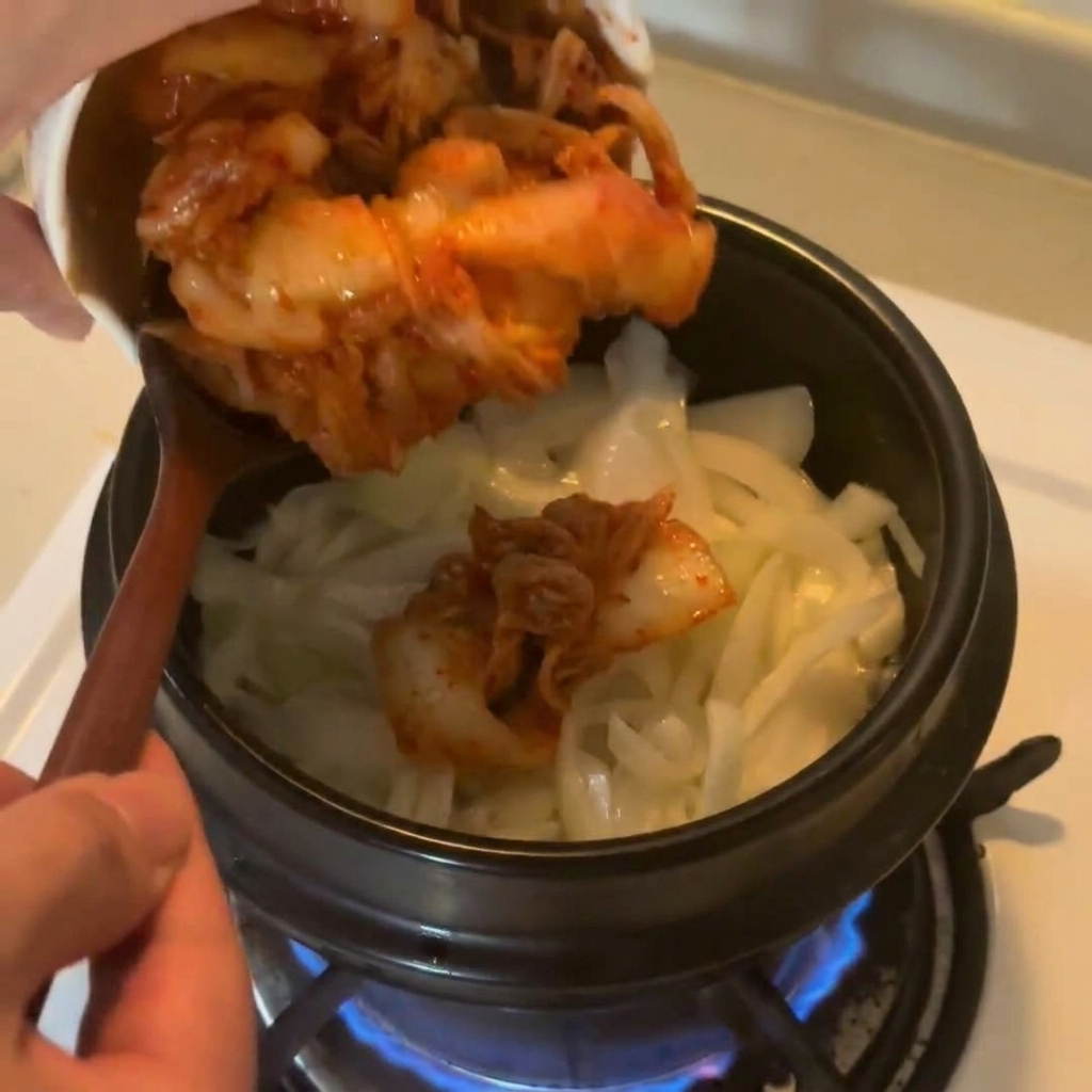 Gehacktes Kimchi wird aus einer weißen Schüssel in den Topf zu den gedünsteten Zwiebeln gegeben.