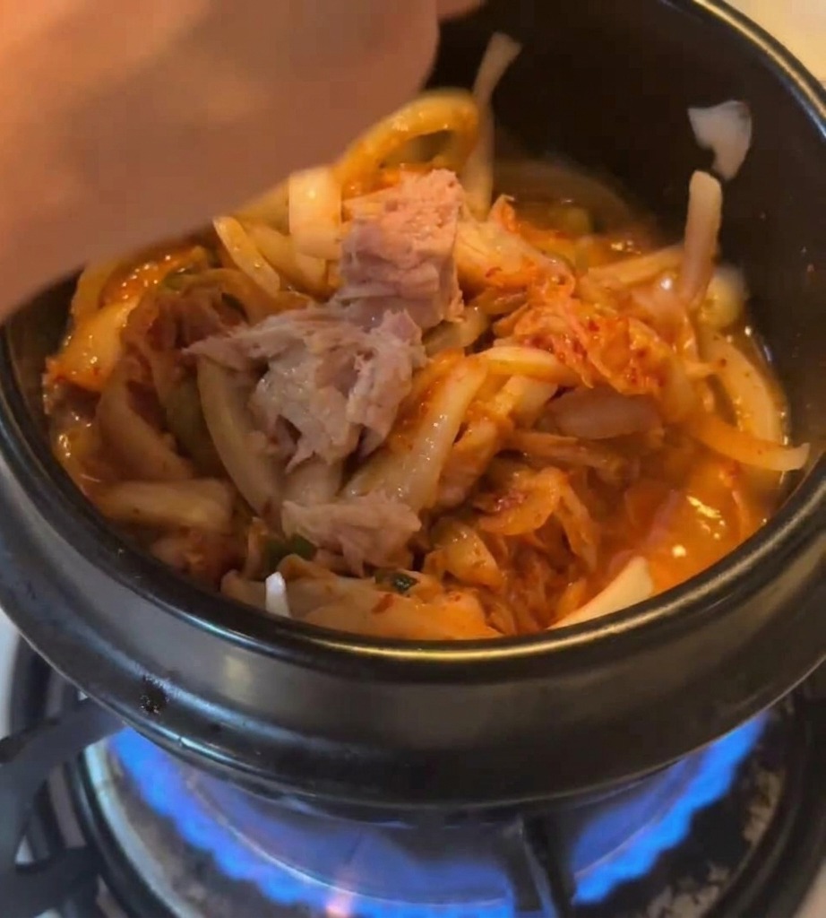 Thunfischstücke werden in den Topf mit Kimchi und Zwiebeln über einer Gasflamme gegeben.