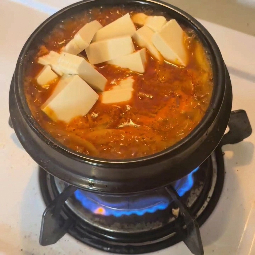 Weiße Tofuwürfel liegen auf dem kochenden, roten Kimchi-Thunfisch-Eintopf im Keramiktopf.