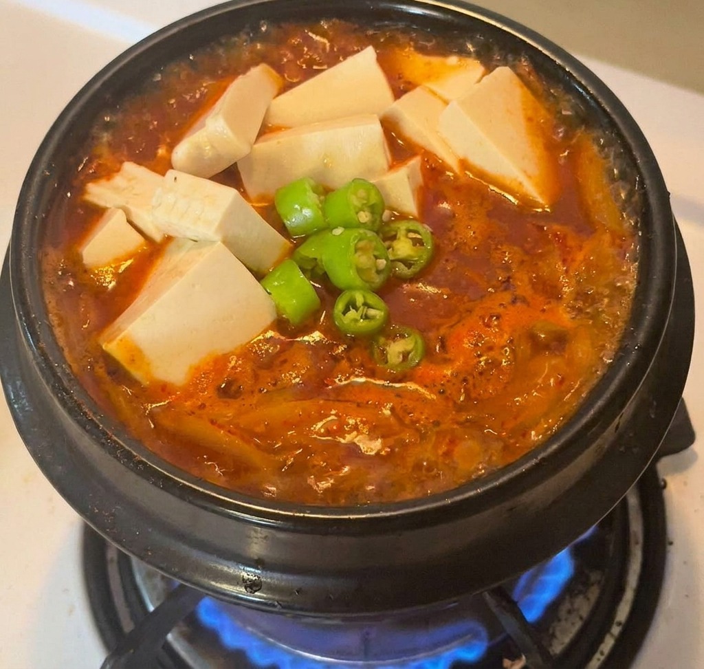 Ein schwarzer Steintopf mit Kimchi-Eintopf, garniert mit Tofuwürfeln und einem Berg grüner Chilischeiben.
