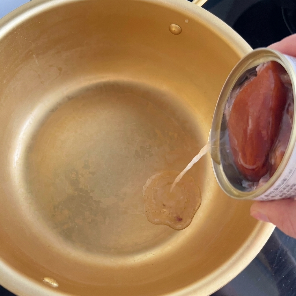 Öl wird aus einer kleinen Dose Thunfisch in die Mitte eines goldfarbenen Kochtopfs gegossen.