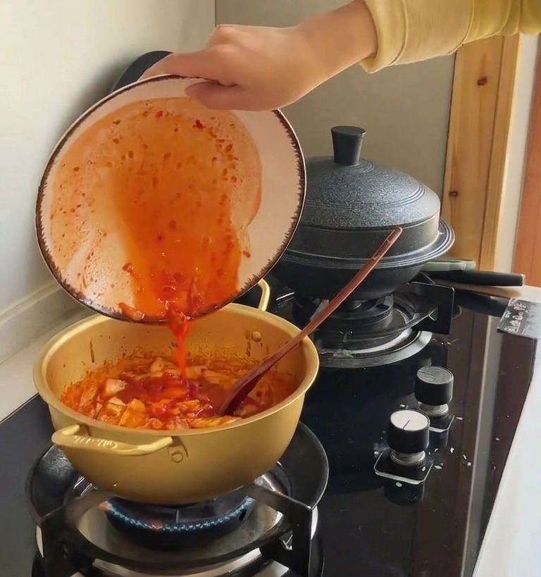 Leuchtend roter Kimchi-Saft wird aus einer Keramikschale in einen goldenen Topf mit gebratenem Kohl gegossen.
