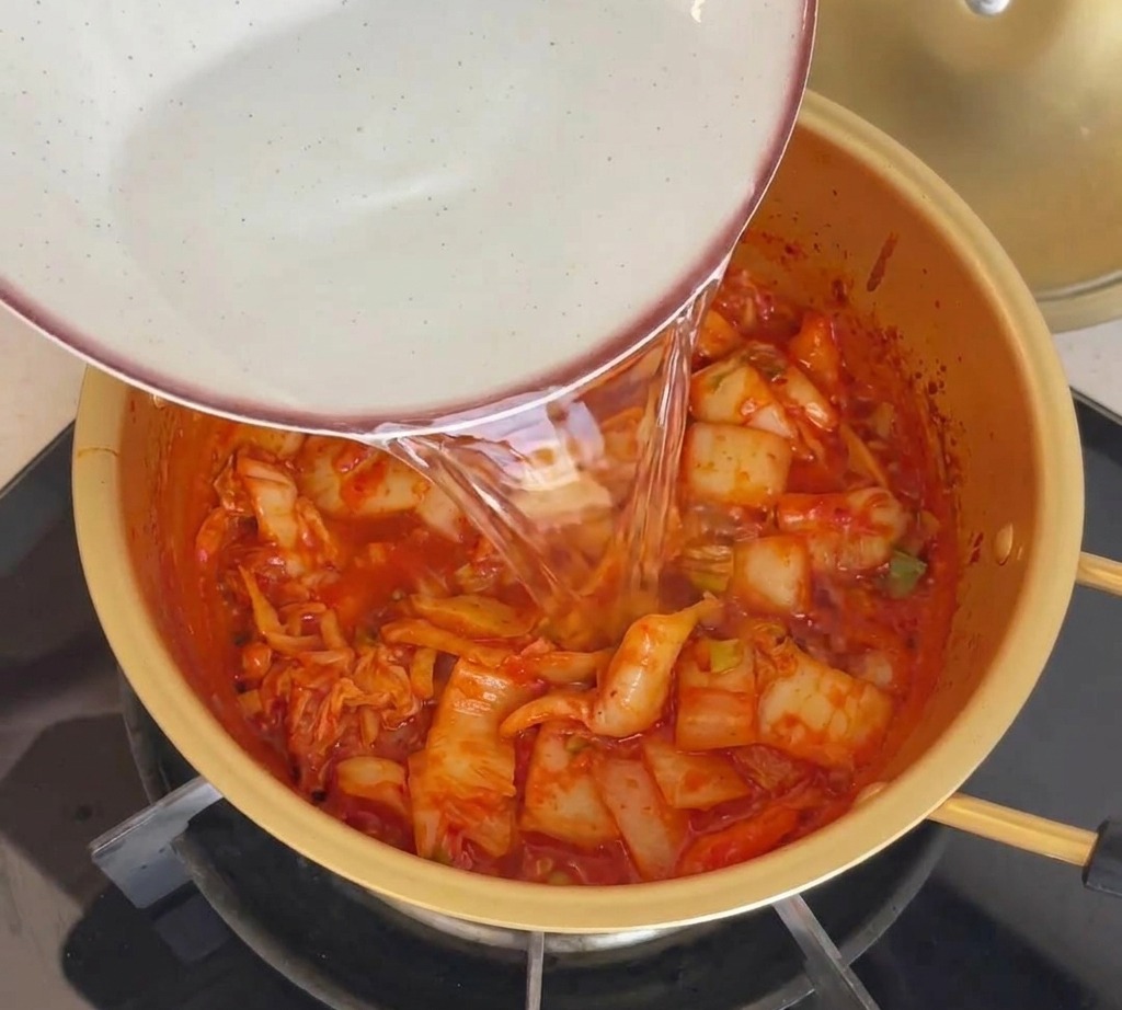 Wasser wird aus einer Keramikschale in einen goldenen Topf mit rotem Kimchi gegossen.
