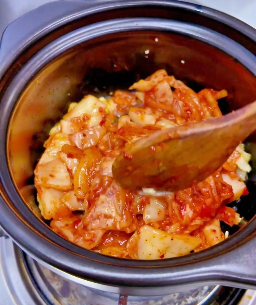 Die Flüssigkeit aus einer Thunfischdose wird über die Kimchi-Knoblauch-Mischung in einen schwarzen Keramiktopf gegossen.