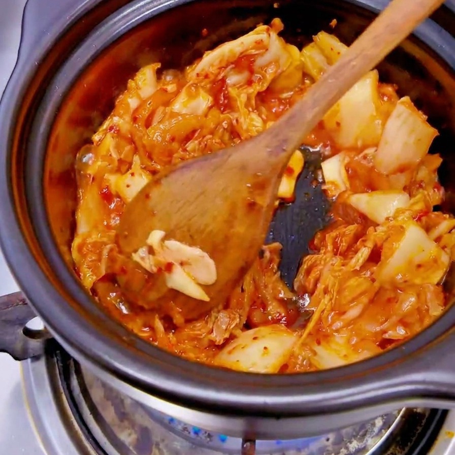 Eine Hand mit schwarzem Handschuh gibt weiße Zwiebelscheiben in einen Topf mit rotem Kimchi.