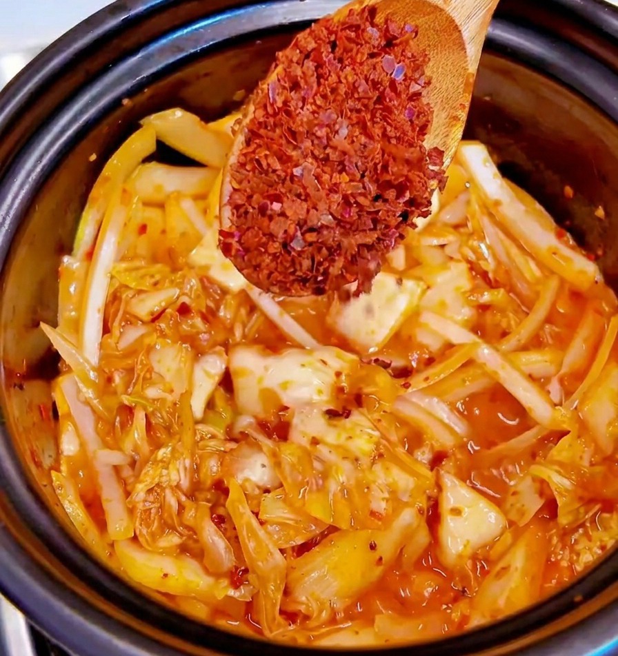 Ein Holzlöffel hält grobe rote Chiliflocken über einen Topf mit Kimchi und Zwiebeln.
