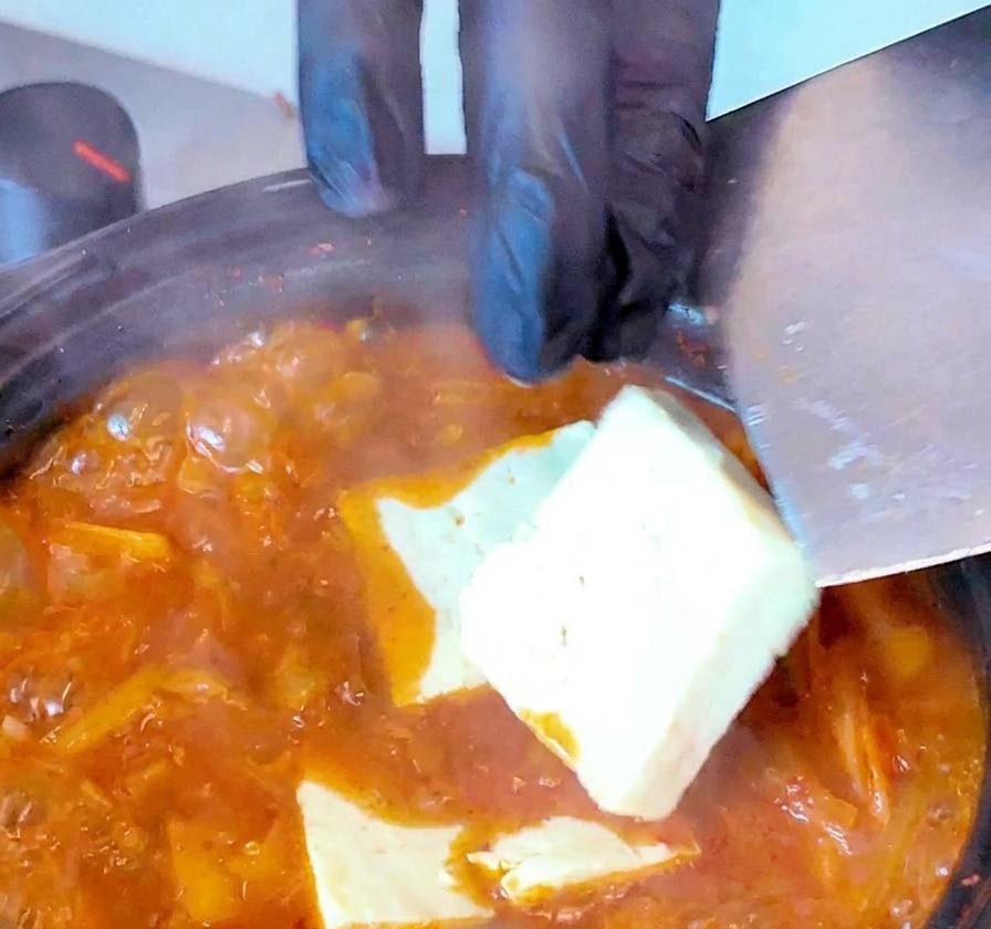 Weiße Tofuscheiben werden vorsichtig mit einem Pfannenwender in die kochende rote Kimchi-Suppe gleiten gelassen.