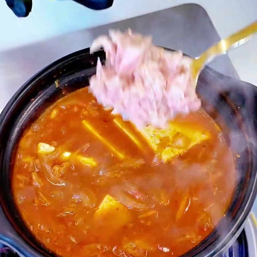 Thunfischstücke werden löffelweise in einen Topf mit kochender Kimchi-Tofu-Suppe gegeben.