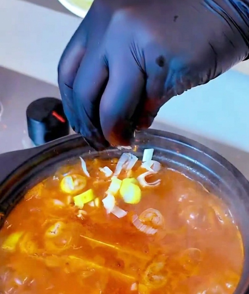 Eine Hand in einem schwarzen Handschuh streut eine Handvoll gehackte Frühlingszwiebeln in einen dampfenden Topf mit rot-oranger Suppe.