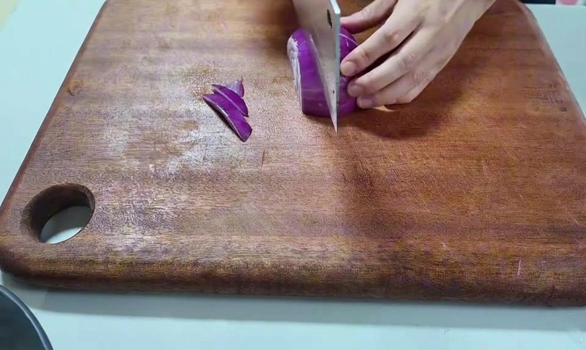 Eine Hand, die eine rote Zwiebel mit einem großen Küchenmesser auf einem Holzbrett schneidet.