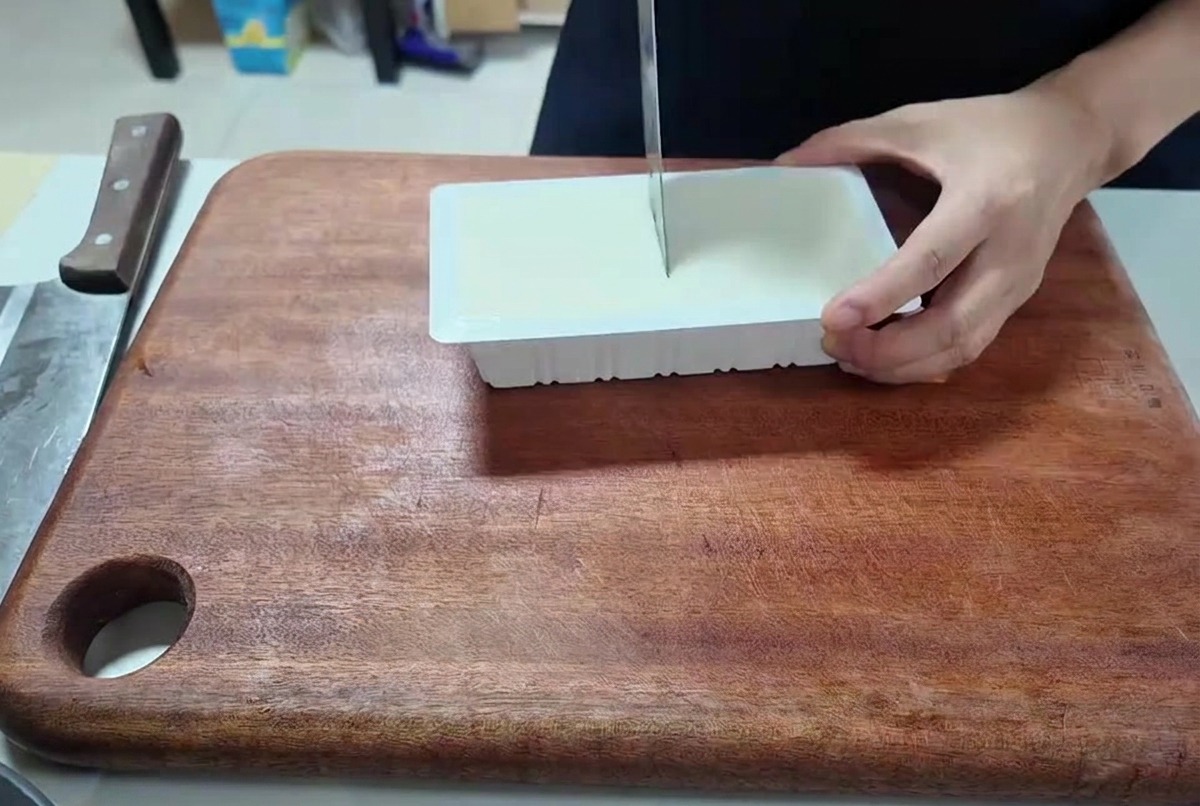 Eine Person schneidet einen Block weißen Tofu mit einem Küchenmesser in gleichmäßige Würfel.