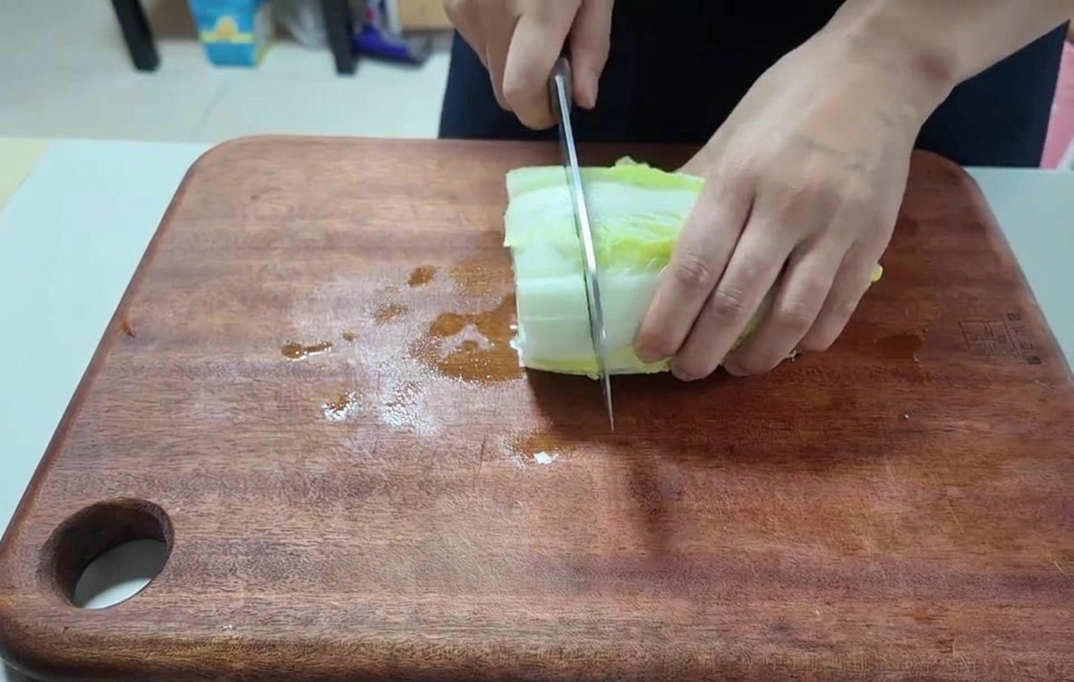 Nahaufnahme von Händen, die frische Chinakohlblätter auf einem dunklen Holzbrett zerkleinern.