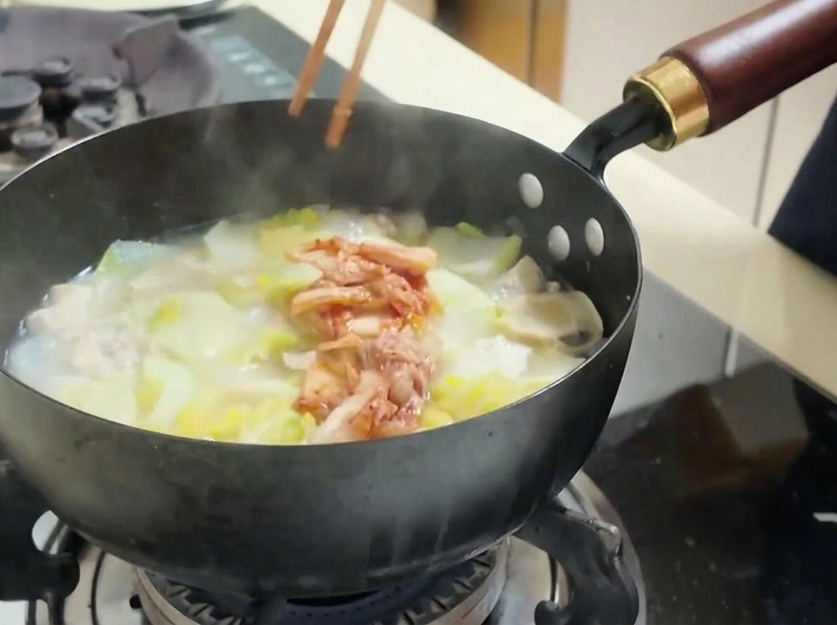 Mit Stäbchen wird rotes, gehacktes Kimchi in den Topf zu Tofu und Kohl gegeben.