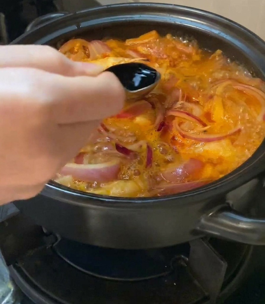 Eine Hand gießt dunkle Sojasauce mit einem Löffel in die köchelnde Kimchi-Suppe.