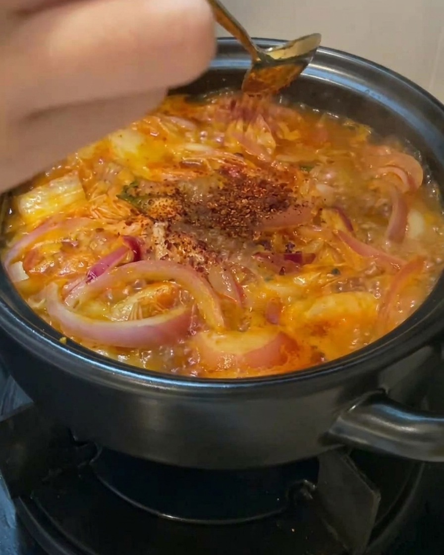 Leuchtend rotes Chilipulver wird mit einem goldenen Löffel über die köchelnde Suppe gestreut.