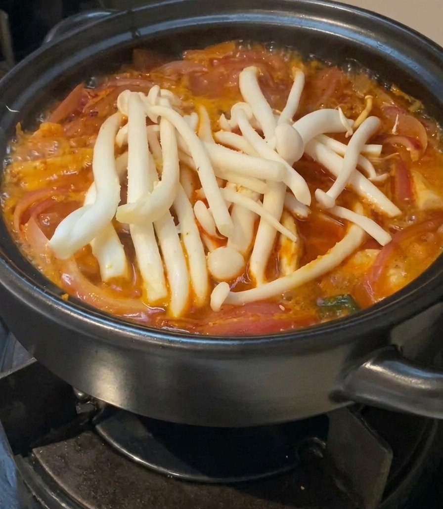 Weiße Buchenpilze liegen auf der Oberfläche der roten, köchelnden Kimchi-Suppe im schwarzen Topf.
