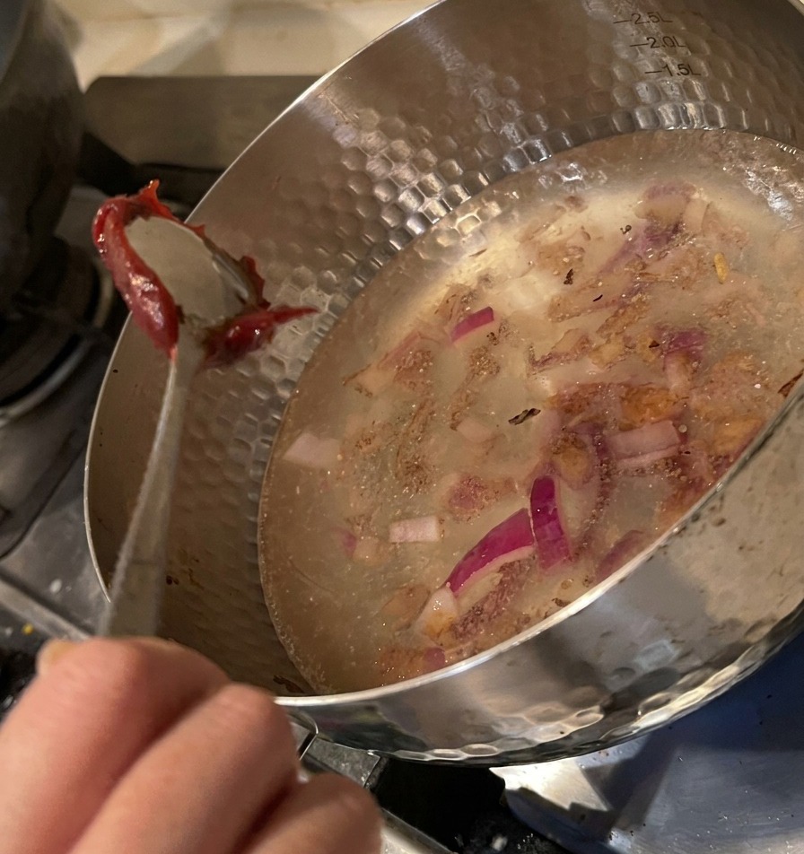 Ein Metalllöffel lässt einen großen Klecks dicke rote Chilipaste in den wassergefüllten Topf mit Zwiebeln gleiten.