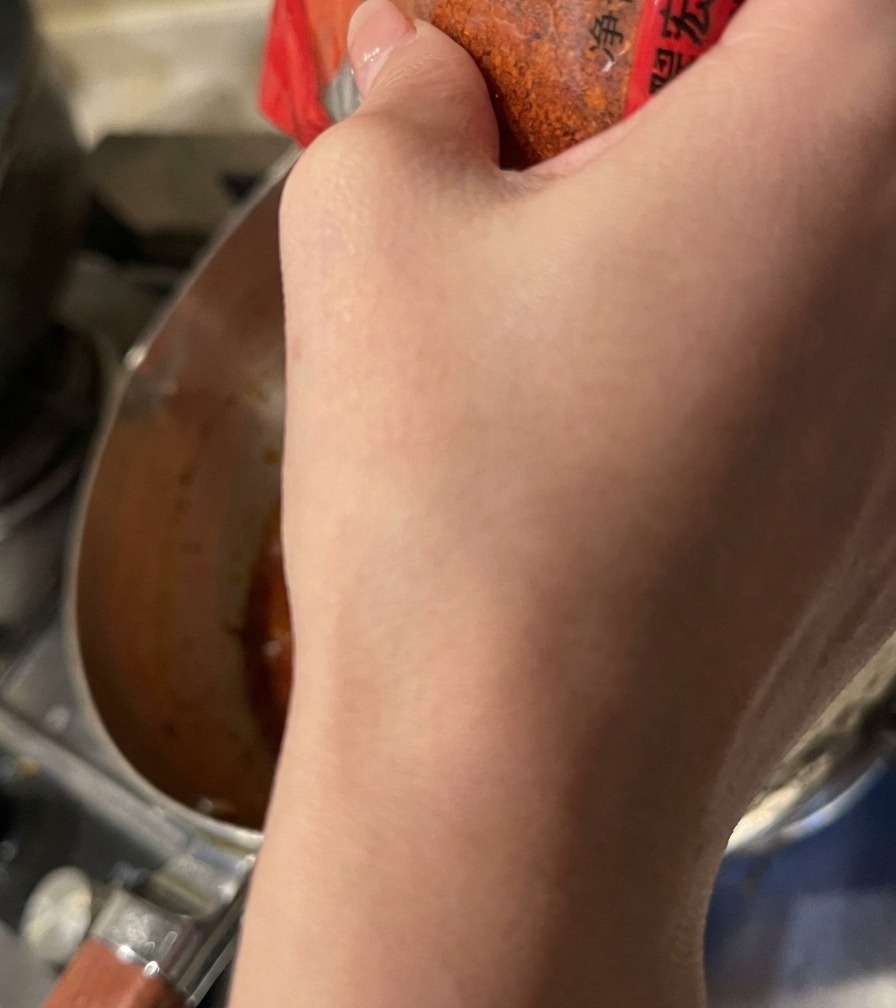 Rotes Chilipulver wird aus einem Behälter in die köchelnde orangefarbene Suppenbasis gestreut.