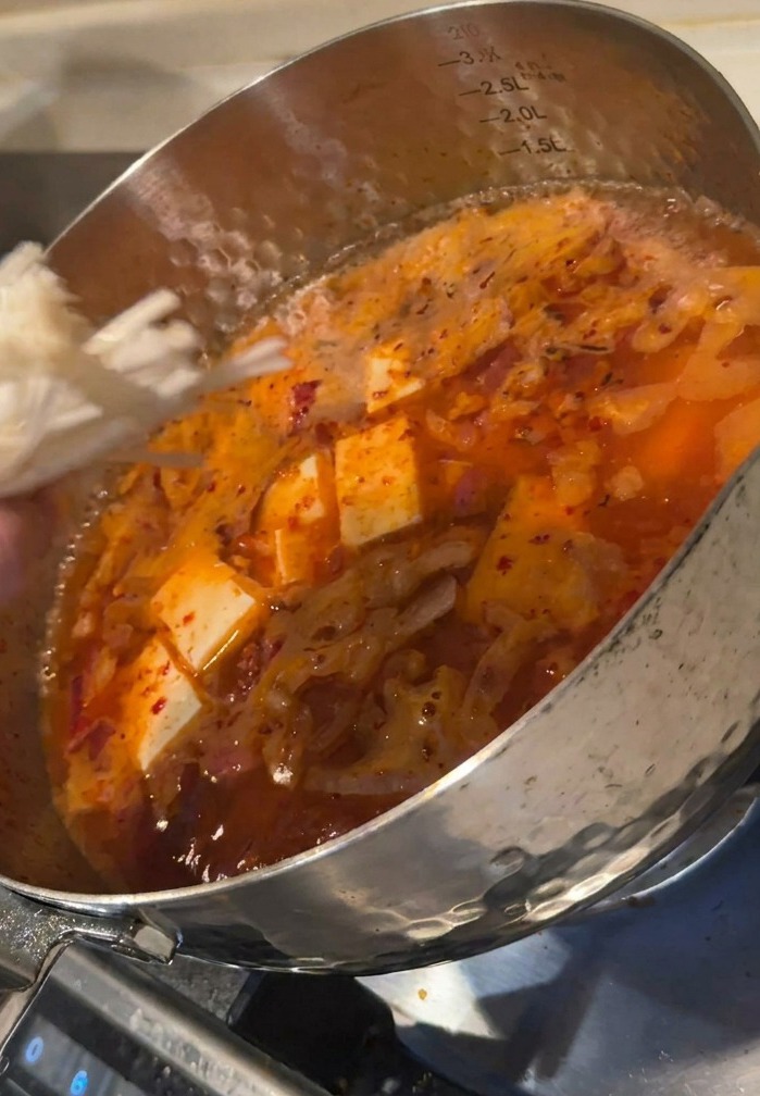 Eine Hand gibt ein Bündel weißer Enoki-Pilze in einen Topf mit sprudelnder roter Kimchi-Suppe und Tofuwürfeln.