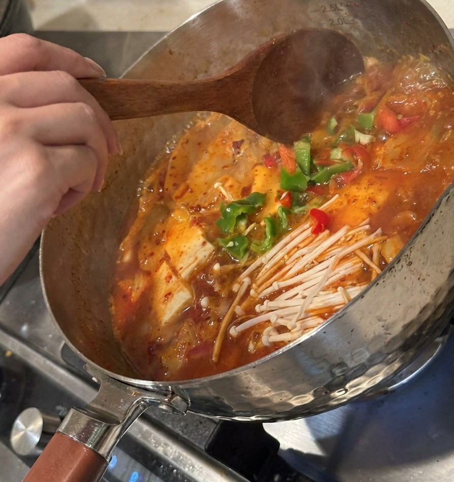 Geschnittene grüne Paprika und fermentiertes Kimchi werden zur Suppe gegeben und mit einem Holzlöffel umgerührt.