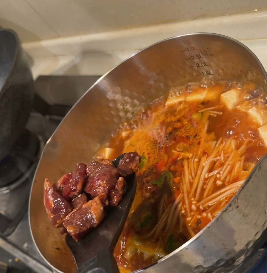 Rohe marinierte Rindfleischwürfel werden mit einem schwarzen Pfannenwender in die kochende rote Gemüsesuppe geschoben.