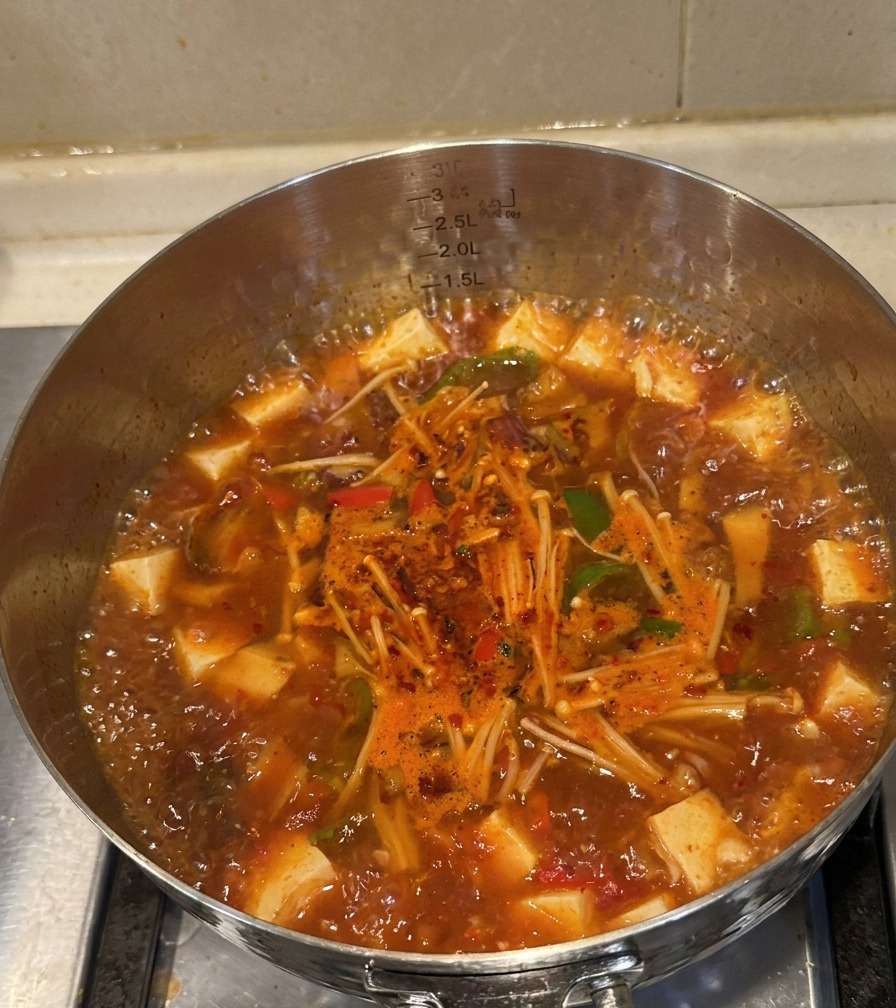 Ein Edelstahltopf mit sprudelnder roter Kimchi-Rindfleisch-Suppe, gegartem Fleisch, Enoki-Pilzen und Paprika.
