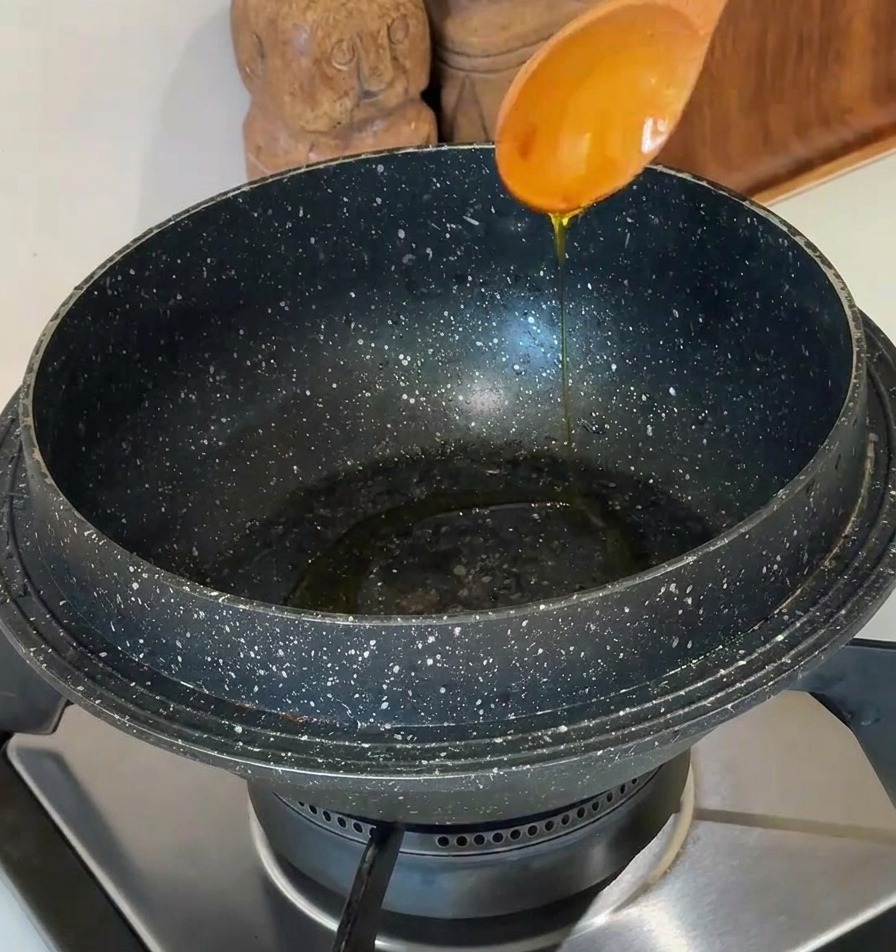 Speiseöl wird aus einem hellen Holzlöffel in einen dunklen Steintopf auf einem modernen Herd gegossen.