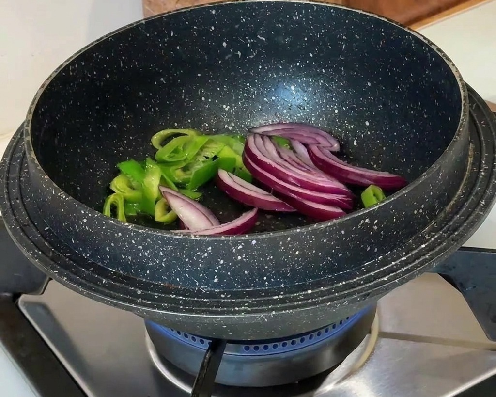 Rote Zwiebelscheiben werden zu den angebratenen Frühlingszwiebeln in einen schwarzen Topf über einer Gasflamme gegeben.