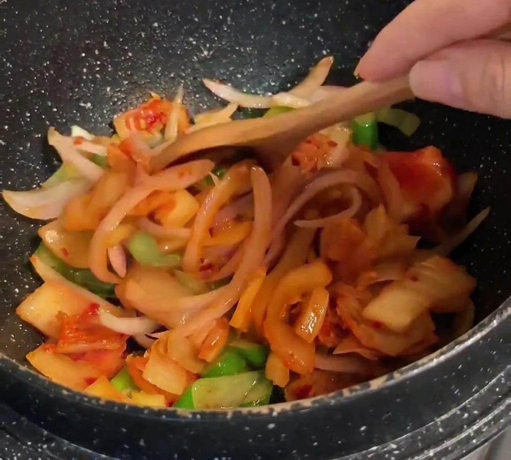 Ein Holzlöffel rührt leuchtend orangerotes Kimchi unter die Mischung aus Zwiebeln und Frühlingszwiebeln.