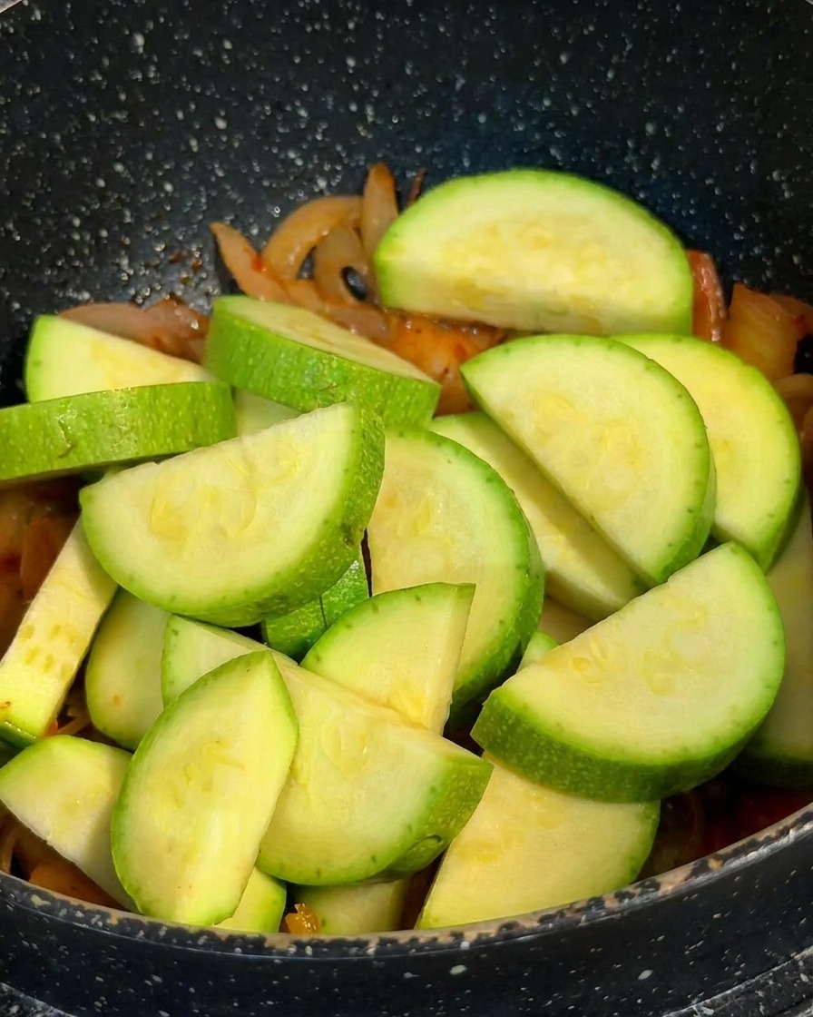 Grüne Zucchini-Halbmonde liegen auf einer Basis aus angebratenem Kimchi und Zwiebeln im Topf.