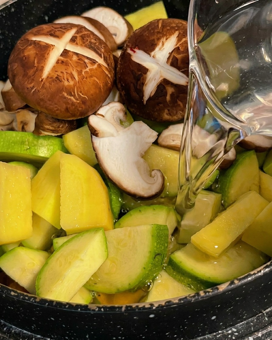 Klares Wasser wird aus einer Karaffe über die Zucchini, Kartoffeln und Shiitake-Pilze im Topf gegossen.