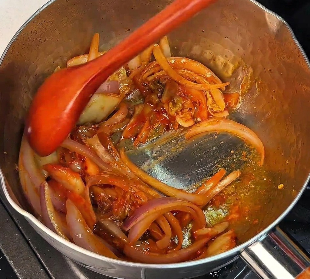 Ein Holzlöffel rührt gehacktes Kimchi und Zwiebeln in einem heißen Metalltopf um.