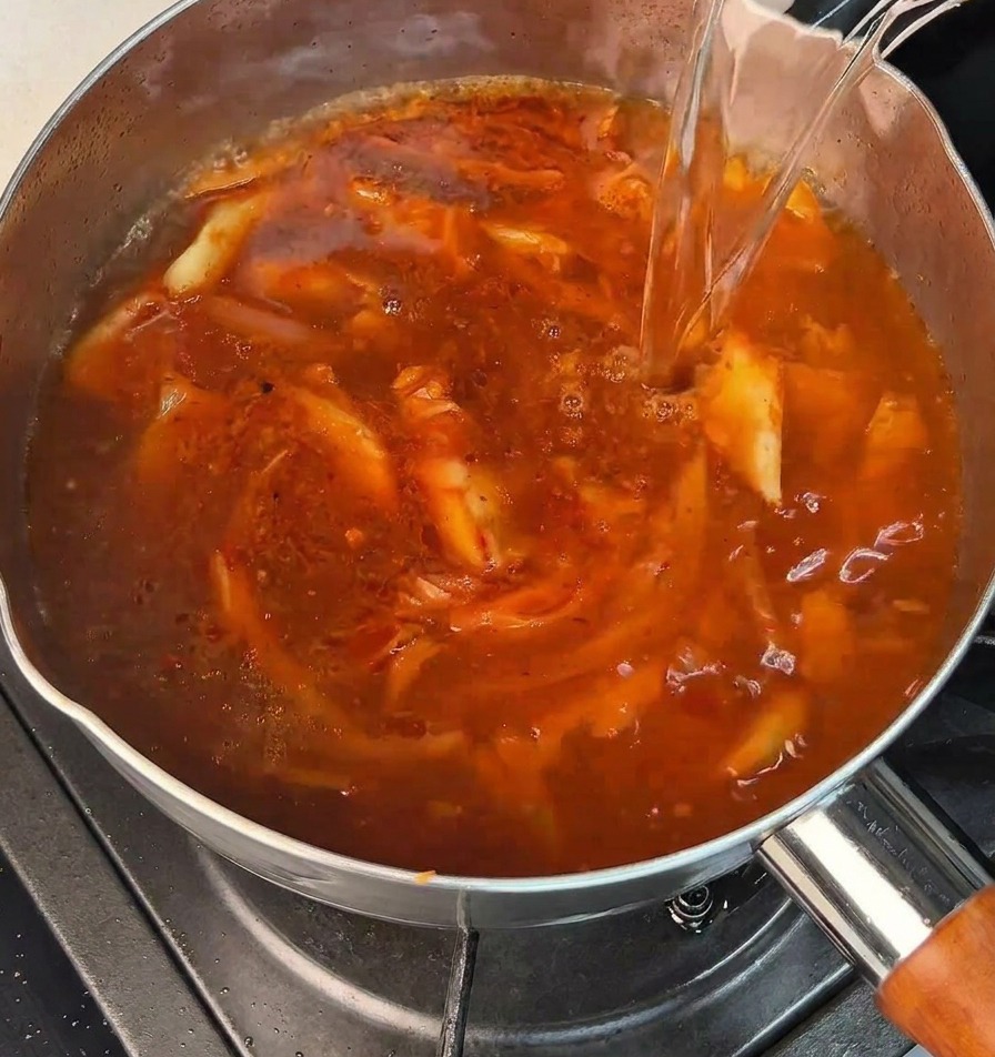 Kochendes Wasser wird in einen Topf mit einer brodelnden, roten Suppenbasis gegossen.
