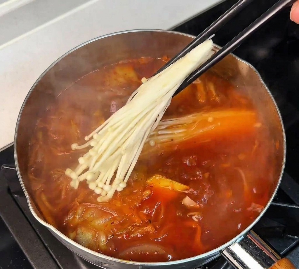 Ein Bündel weißer Enoki-Pilze wird mit Stäbchen in die köchelnde Kimchi-Suppe gelegt.