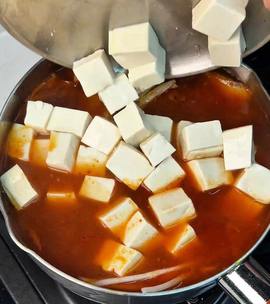 Weiße Tofuwürfel werden aus einer Edelstahlschüssel in die Kimchi-Brühe gegeben.