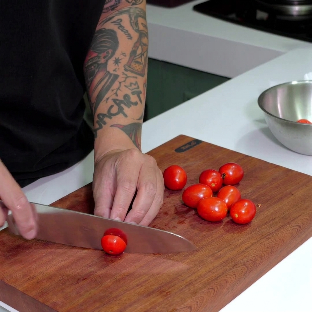 Ein Kochmesser schneidet leuchtend rote Kirschtomaten auf einem dunklen Holzschneidebrett.