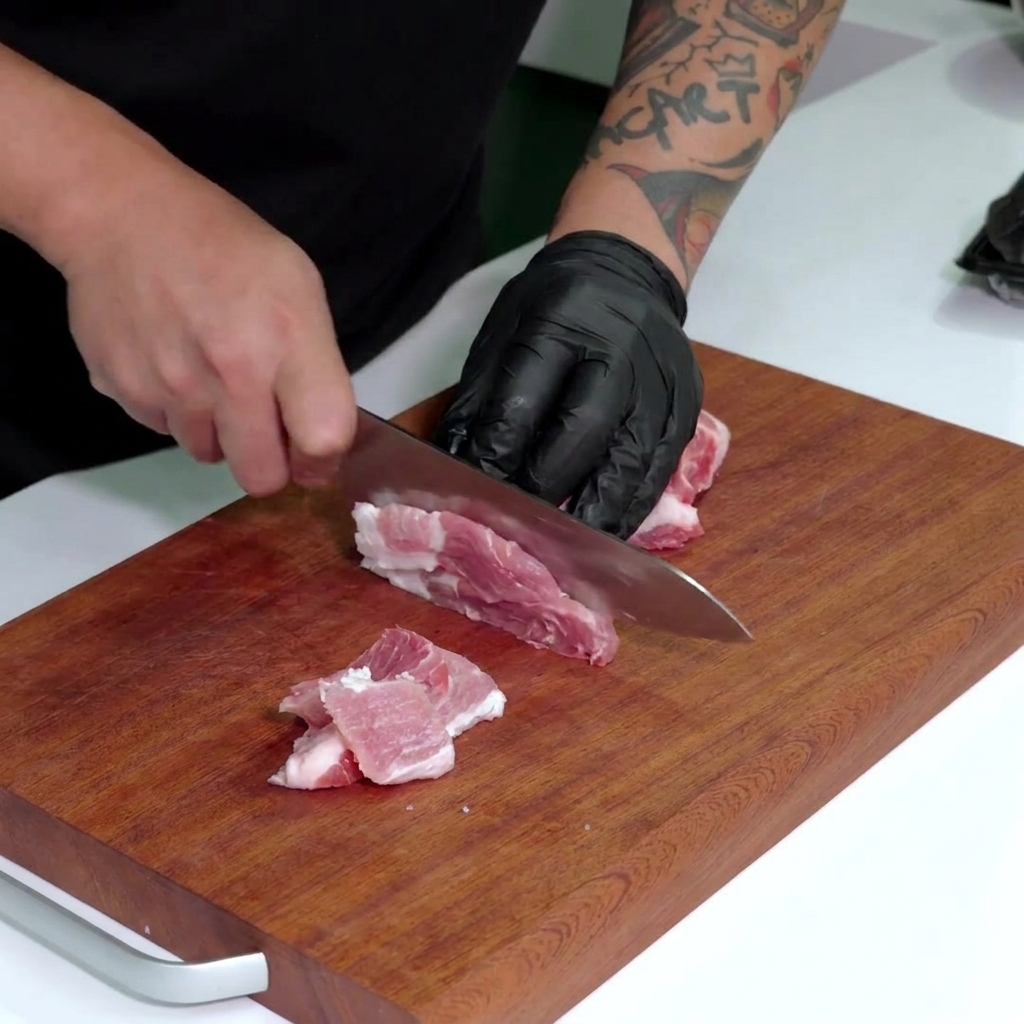 Eine Hand in einem schwarzen Handschuh schneidet rohen Schweinebauch in schmale Streifen.