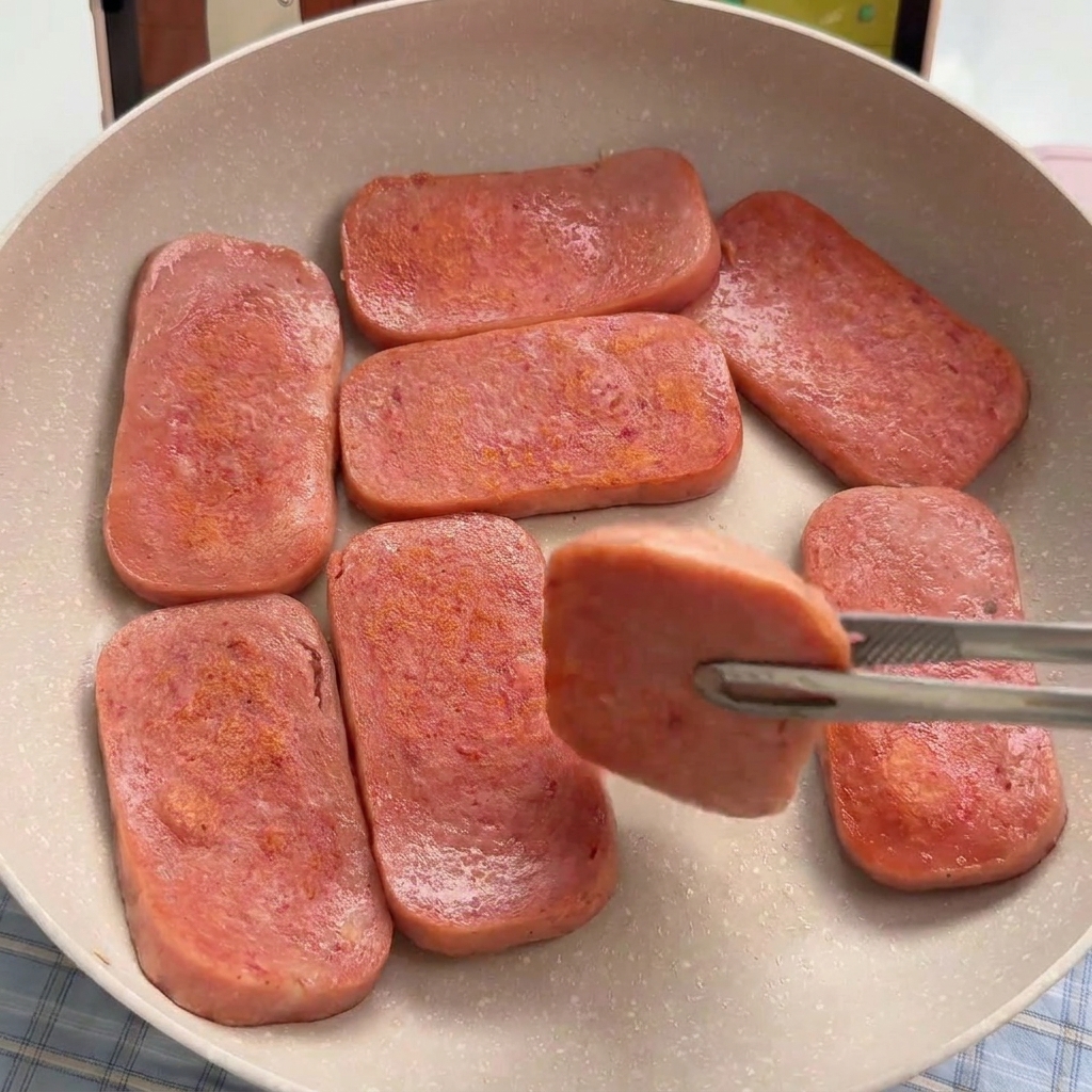 Rechteckige Scheiben von rosa Frühstücksfleisch werden in einer hellen Pfanne goldbraun angebraten.