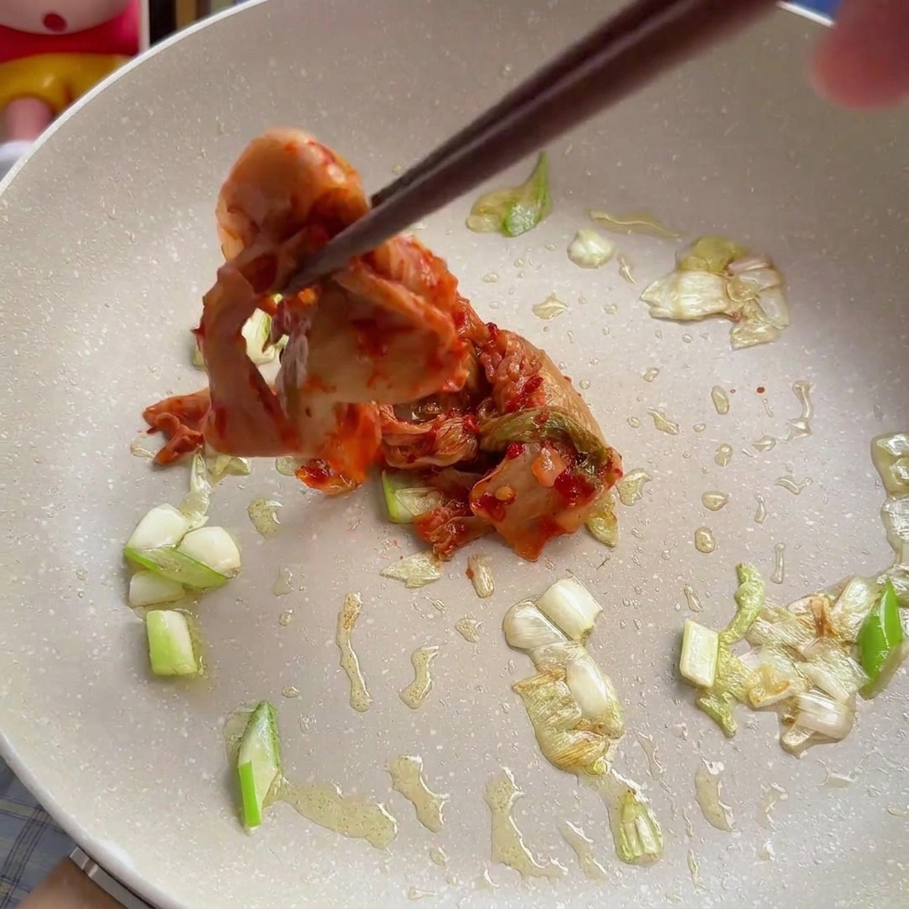 Mit Stäbchen wird eine Portion rotes Chinakohl-Kimchi zu den gedünsteten Zwiebeln in die Pfanne gegeben.