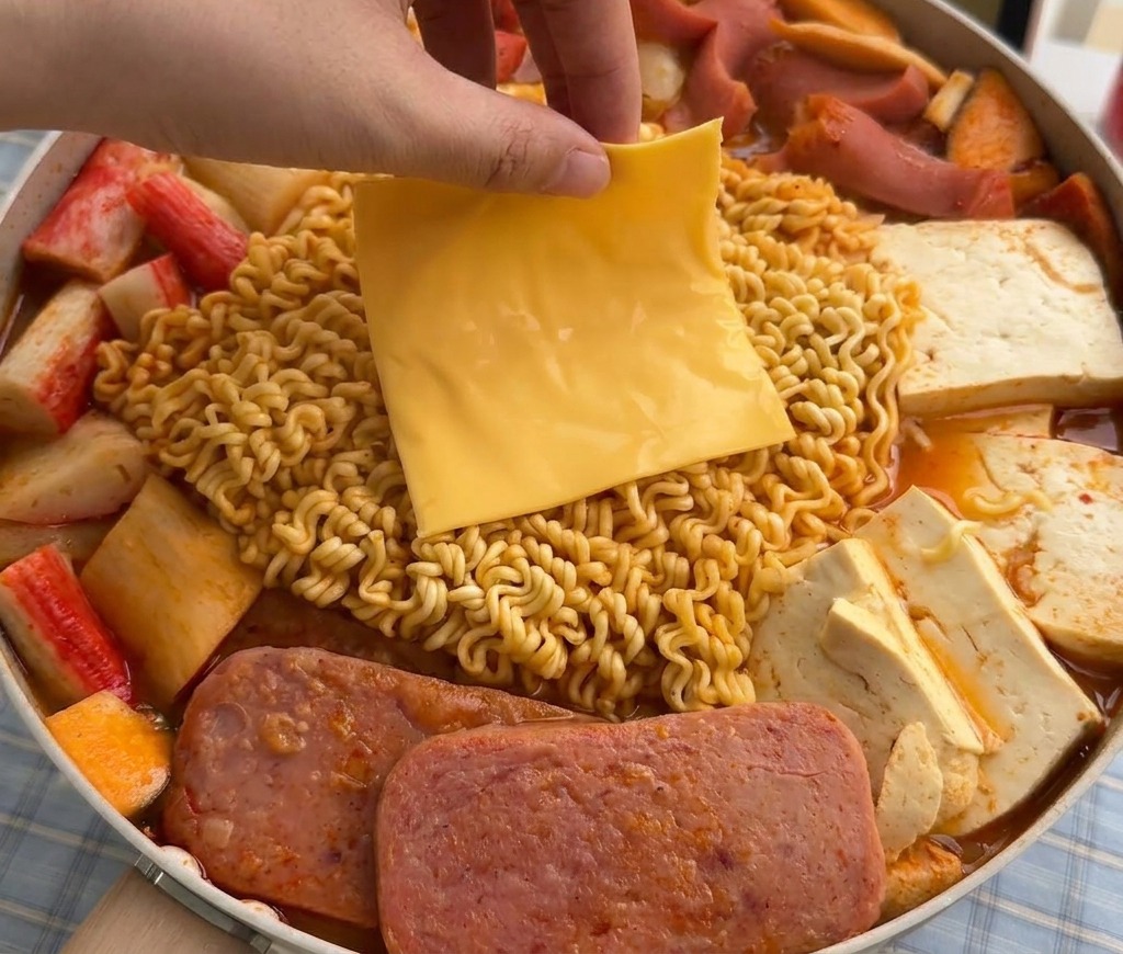 Eine Hand legt eine Scheibe gelben Käse auf die weich werdenden Ramen-Nudeln im dampfenden Eintopf.