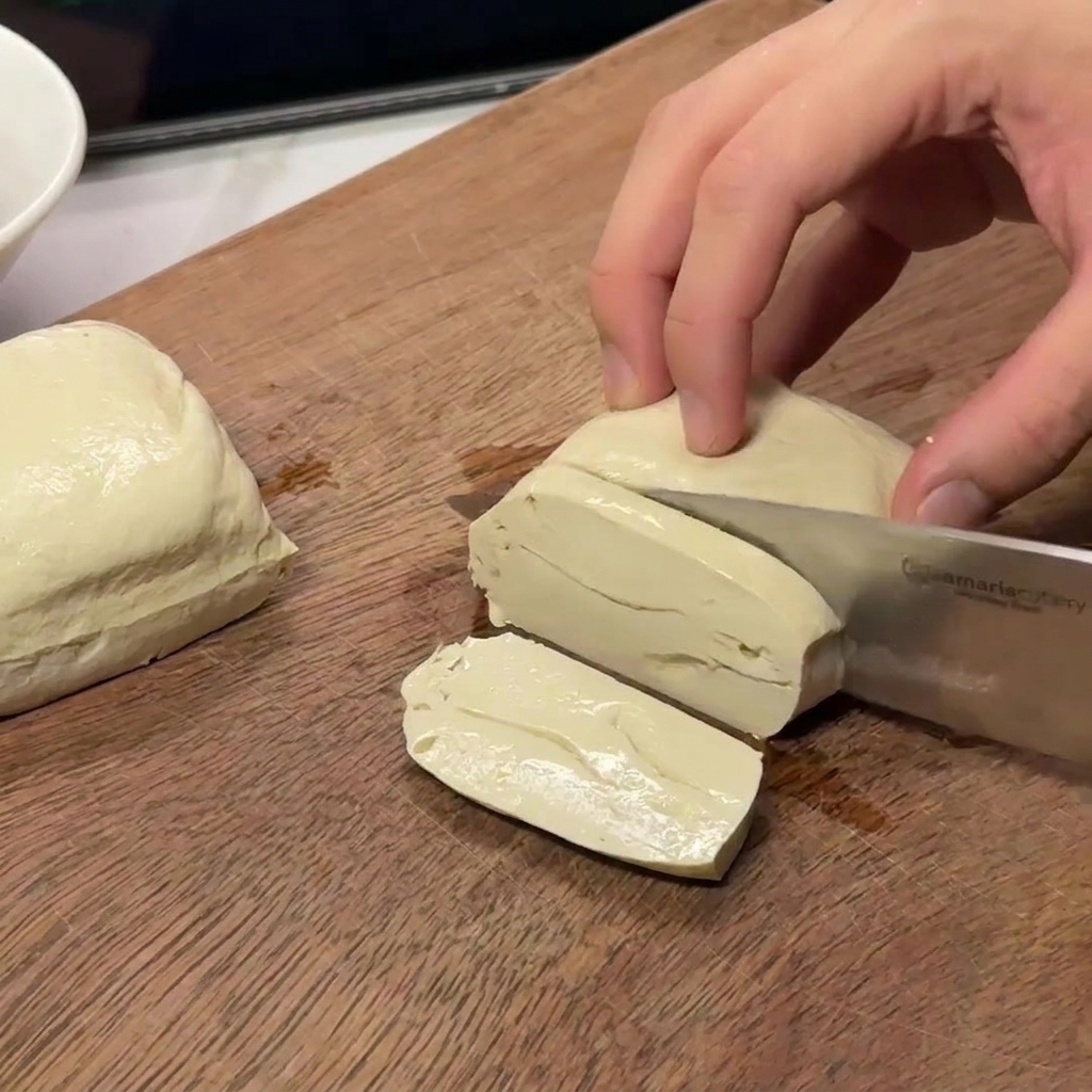 Hände schneiden einen Block festen weißen Tofu in gleichmäßige rechteckige Stücke auf einer Holzoberfläche.