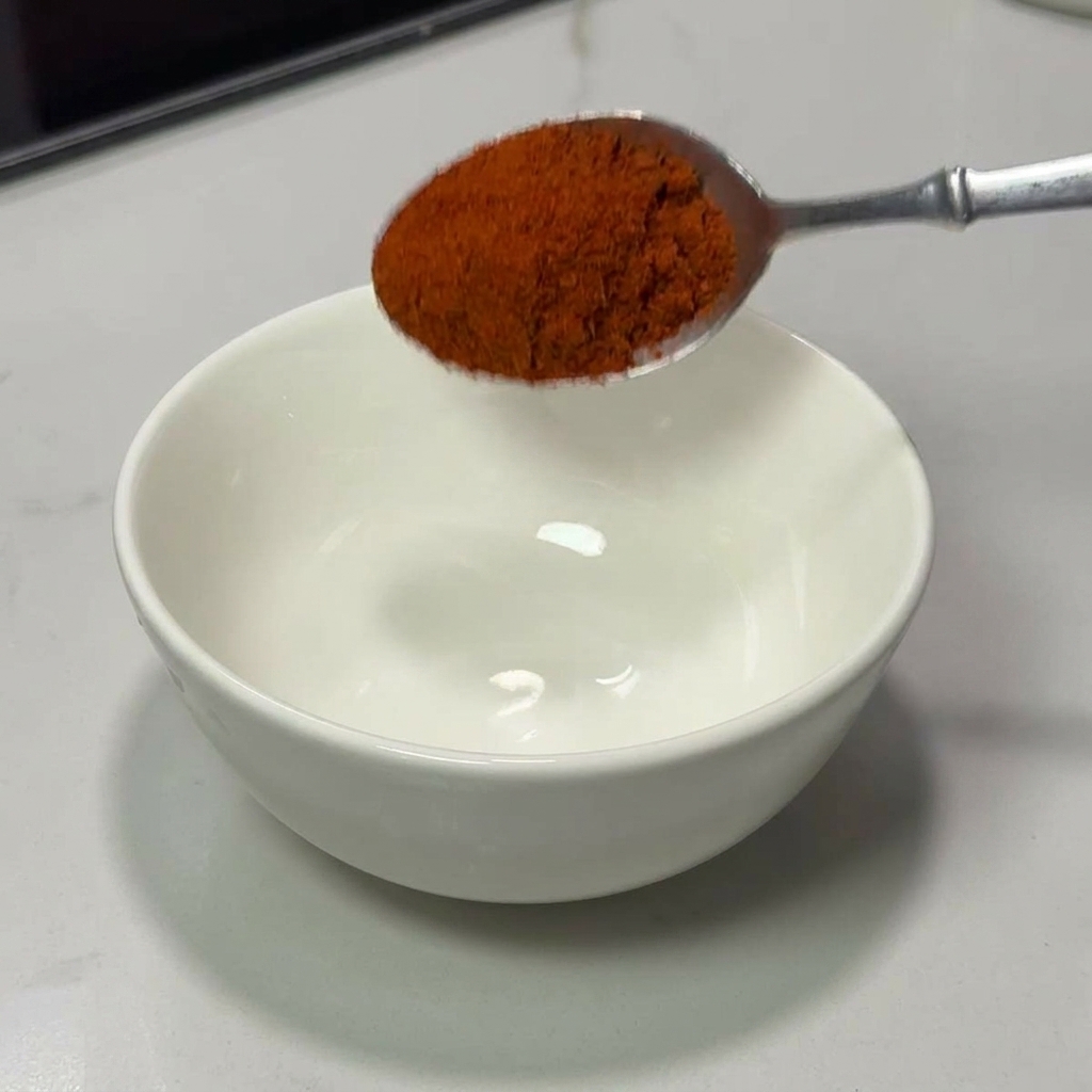 Ein Silberlöffel gießt leuchtend rotes Chilipulver in eine weiße Keramikschale.