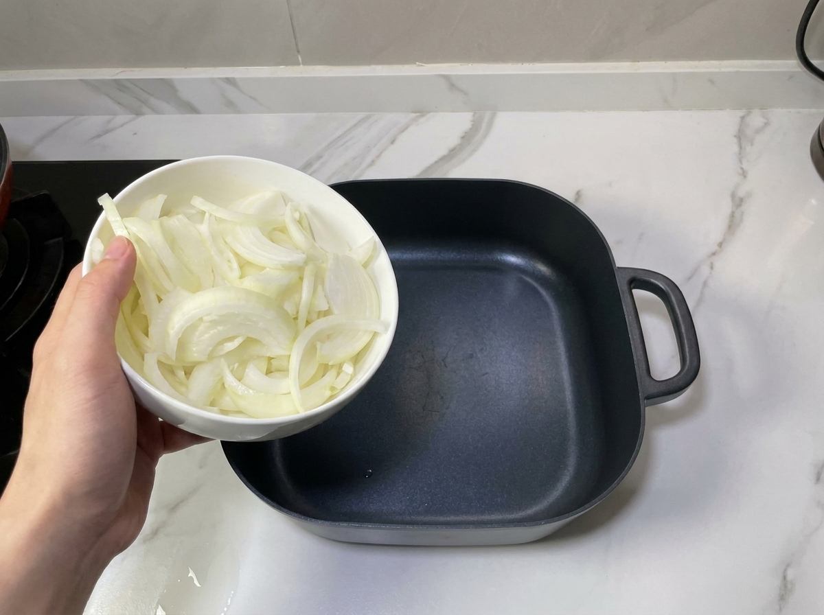 Eine Hand hält eine Schüssel mit Zwiebelringen über einen leeren schwarzen Kochtopf zum Schichten.