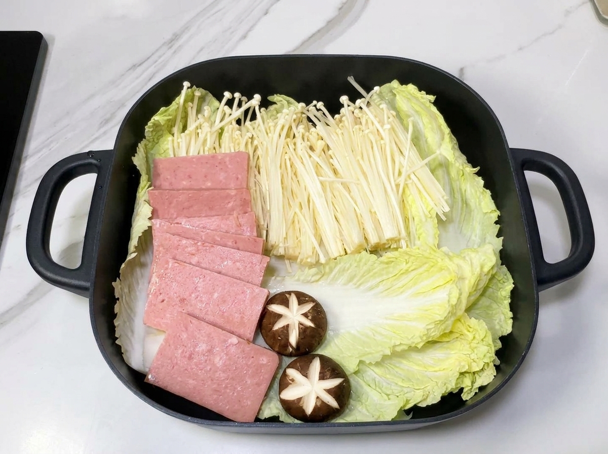 Ein quadratischer Topf mit Schichten aus Chinakohl, Enoki-Pilzen und Spam-Scheiben auf einem Zwiebelbett.