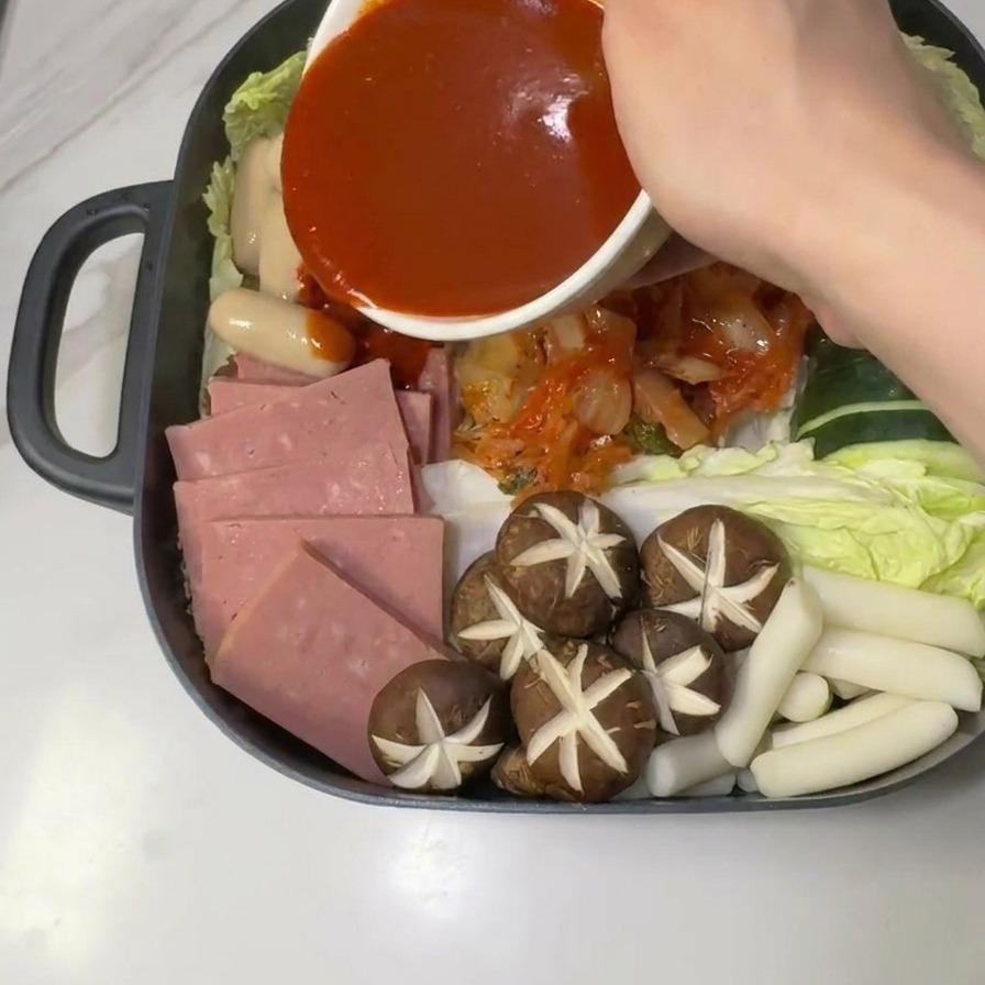 Scharfe rote Gochujang-Sauce wird aus einer weißen Schüssel mittig über die Zutaten des Army Stews gegossen.